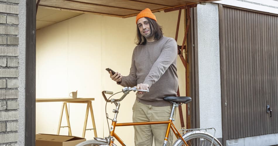 Man met een oranje muts staat voor een garage met een fiets en een smartphone
